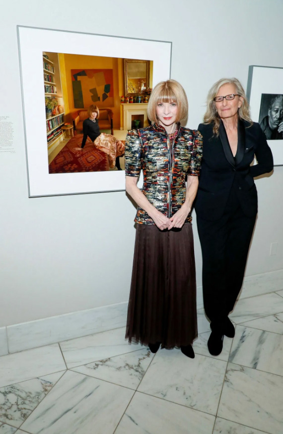 woman in a long dress and another woman in black pants in front of a photo