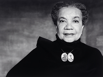 Bust length photo of a Black woman wearing a black wrap and portrait jewelry of abolitionists