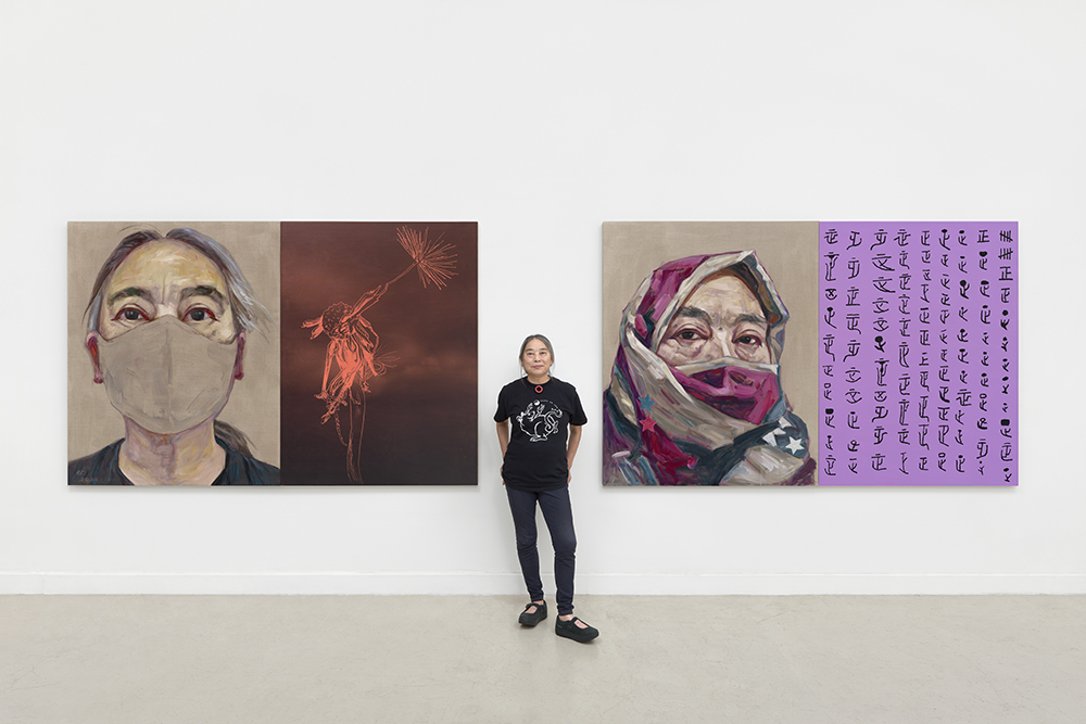 Woman wearing black stands between two enormous self portraits of herself wearing a mask