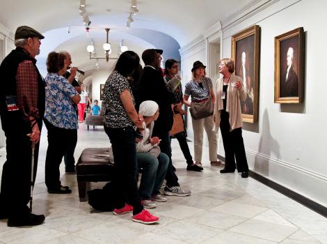 A group of people gathered around a portrait listen to a woman pointing to the wall