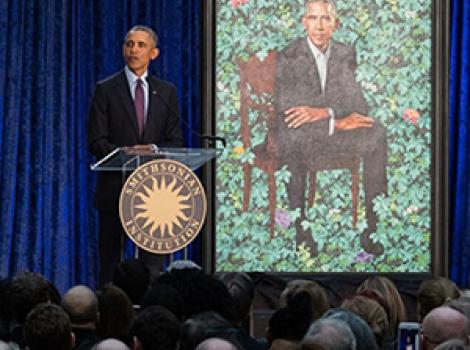 A man (Barack Obama) speaking at a podium in front of his portrait