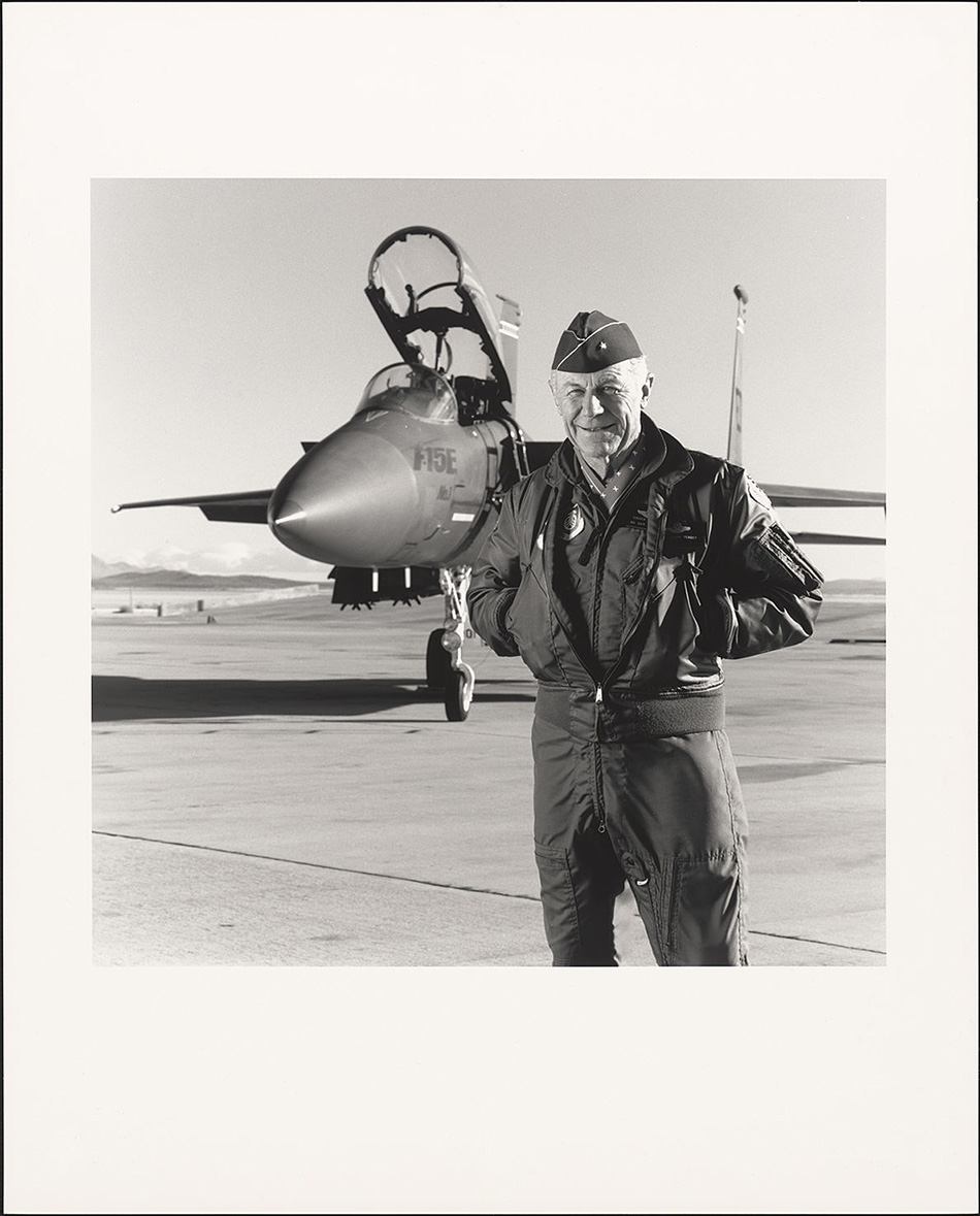 older man in a flight suit standing in front of a jet plane